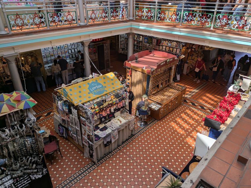 Interior view of a busy Camden Market stall area, showcasing an upper and lower level with decorative railings and multiple vendors. The lower level features a vendor stall with a yellow and blue sign labeled 'London T-Shirts,' displaying clothing and accessories on display racks and tables, attended by staff. The surrounding floor is tiled with small, reddish-brown and black patterned tiles, appearing clean and well-maintained. To the left, a display of jewelry and souvenirs is partially visible, with a multicolored umbrella providing shade. The upper level overlooks the bustling market scene below, illuminated by natural light filtering through the openings. This scene highlights the importance of surface cleaning, organisation, and hygiene practices essential for maintaining a tidy and inviting shopping environment, as promoted by Camden Carpet Cleaning.
