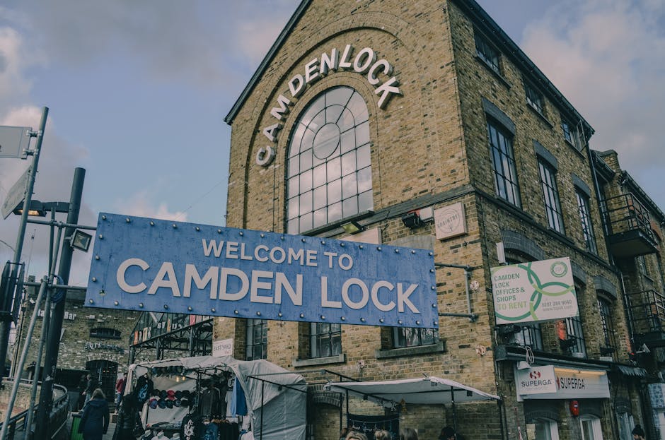 Exterior view of a brick building with large arched windows and a prominent circular window at the top, displaying the words 'Camden Lock' in white letters. A blue signboard with white text reading 'Welcome to Camden Lock' is mounted on a metal fence in front of the building. Below the sign, there are small market stalls selling clothing and accessories, sheltered under white canopies. The building features a modern industrial design with black window frames, metal balcony railings, and brickwork in earthy tones. The scene is illuminated by natural daylight with partly cloudy skies overhead, creating a well-lit, clean appearance. Camden Carpet Cleaning logo is not visible in the image.