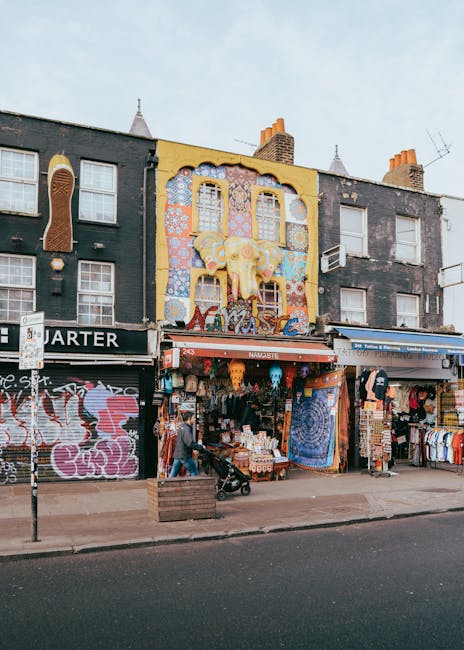 Interior view of a busy Camden Market stall area, showcasing an upper and lower level with decorative railings and multiple vendors. The lower level features a vendor stall with a yellow and blue sign labeled 'London T-Shirts,' displaying clothing and accessories on display racks and tables, attended by staff. The surrounding floor is tiled with small, reddish-brown and black patterned tiles, appearing clean and well-maintained. To the left, a display of jewelry and souvenirs is partially visible, with a multicolored umbrella providing shade. The upper level overlooks the bustling market scene below, illuminated by natural light filtering through the openings. This scene highlights the importance of surface cleaning, organisation, and hygiene practices essential for maintaining a tidy and inviting shopping environment, as promoted by Camden Carpet Cleaning.