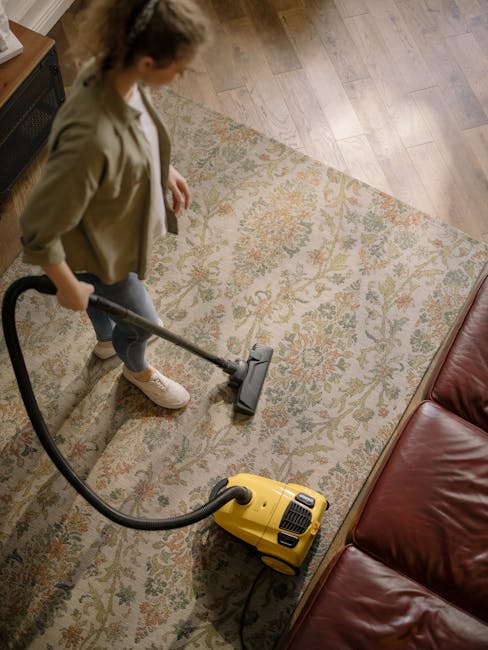 A person performing surface cleaning on a patterned area rug using a yellow vacuum cleaner with a black hose, in a living room with wooden flooring and a leather sofa positioned nearby. The room is well-lit, showcasing the clean and dust-free condition of the rug and surrounding surfaces. The individual is dressed casually, focusing on deep cleaning to maintain hygiene and freshness, with visible tools indicating professional domestic cleaning services provided by Camden Carpet Cleaning for Chalk Farm NW1 flats.