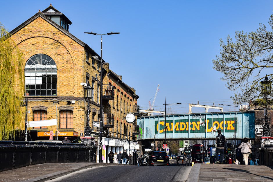 A large, weathered metal railway bridge painted in faded turquoise with bold yellow letters spelling 'CAMDEN LOCK' and a small illustration of a person hanging from a rope. The underside of the bridge reveals visible rust and peeling paint. Below, a busy street scene features asphalt roads with painted white markings, pedestrian crossings, traffic lights, and sidewalks with pedestrians walking in various directions. The background includes buildings with brick facades and some greenery, under an overcast sky that gives the scene a muted, subdued lighting. This urban setting exemplifies exterior cityscape features relevant to Camden in North London, highlighting street-level activity and infrastructural elements.