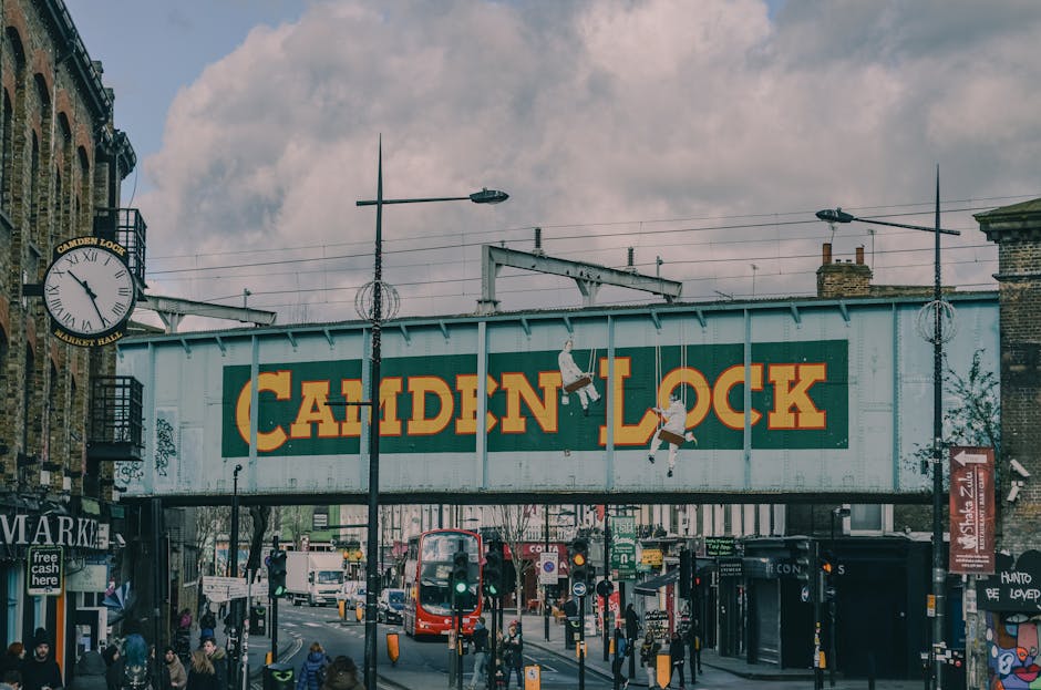 A street scene near Camden Town station in London during daytime, featuring the prominent Camden Lock sign on a large, weathered metal structure spanning across the road. The sign displays bold yellow and red lettering on a green background. Surrounding the sign are classic brick buildings, some with shopfronts and signage, including a clock on the left side showing approximately 12:25. The street is busy with pedestrians walking on the sidewalk, some waiting at traffic lights, and various vehicles such as a red double-decker bus, cars, and bicycles moving through the intersection. The scene is well-lit with natural daylight, and the sky is partly cloudy, casting soft shadows. Utility poles with streetlights and overhead wires are visible, capturing an active, urban environment near Camden Town station, an area serviced by Camden Carpet Cleaning for domestic and commercial surface cleaning needs.