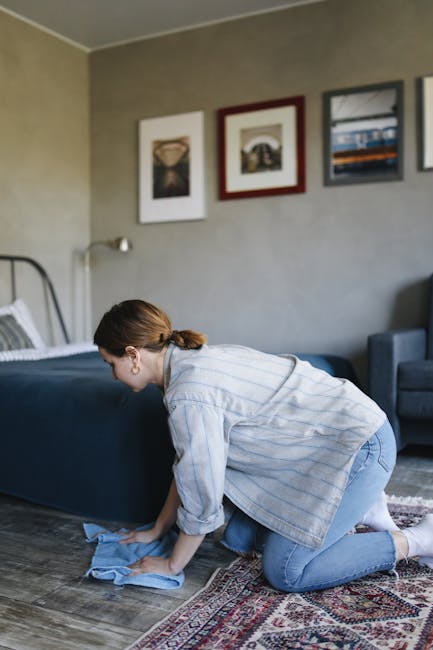 A person kneeling on a patterned carpet in a room, inserting a cloth into a yellow and black vacuum cleaner. The scene shows part of the person's arm and leg, with the carpet displaying an intricate floral design in shades of cream, green, and brown. The vacuum cleaner, positioned on the carpet, has a large hose attached, and the open lid reveals storage space inside. The room is well-lit, emphasizing the cleanliness of the carpet and the presence of cleaning tools, illustrating a domestic cleaning process. This image relates to surface cleaning and deep cleaning, consistent with Camden Carpet Cleaning's services discussed on the page titled 'Same day carpet cleaning Camden what to know about delays, CAMDEN' at camdencarpetcleaning.co.uk.